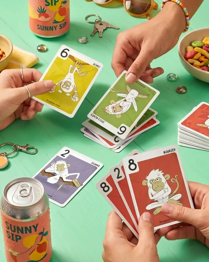Manos emparejando cartas de mono del mismo color sobre una mesa verde, con refrescos y aperitivos alrededor